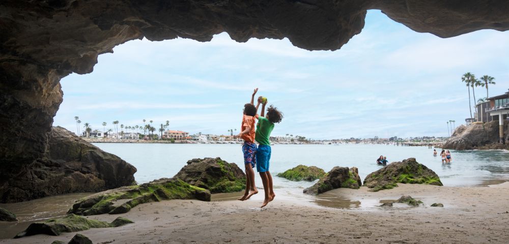 Two children joyfully jump near a sandy beach, framed by a cave, with palm trees and houses visible across the water.