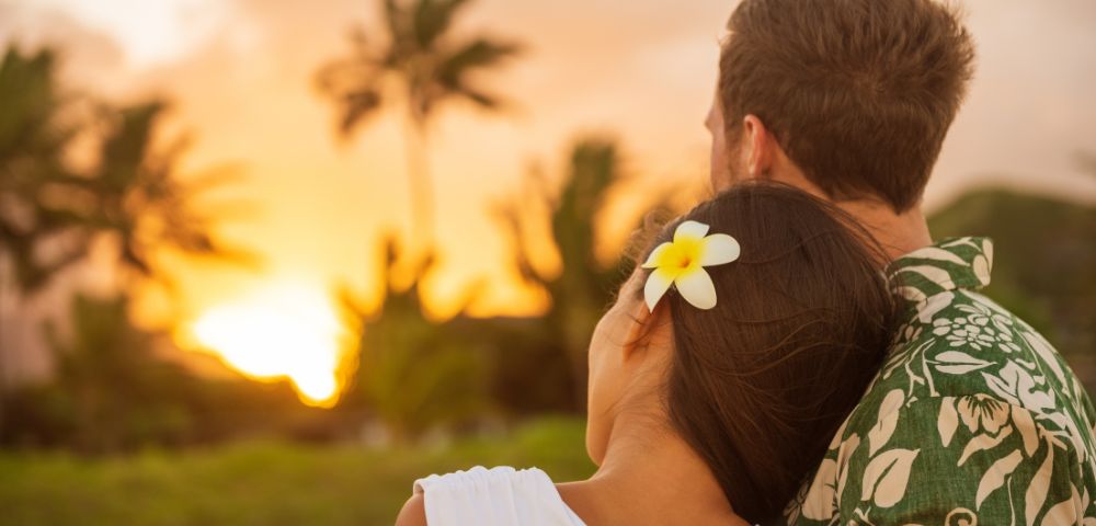 A couple gazes at a vibrant sunset, standing close together. The woman rests her head on the man's shoulder, wearing a white flower in her hair. Palms silhouette against the colorful sky, evoking a serene, romantic atmosphere.