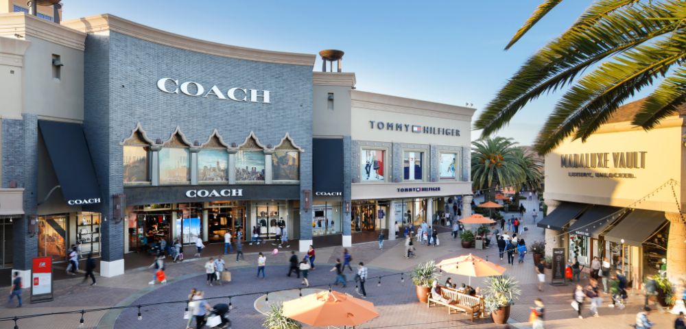 A lively outdoor shopping area features Coach, Tommy Hilfiger, and Mandalay Vault stores. Shoppers and palm trees create a vibrant, sunny atmosphere.