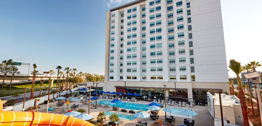 Modern hotel exterior with a tall white building, pool area, and blue-striped umbrellas. Sunny, relaxed atmosphere with surrounding palm trees.