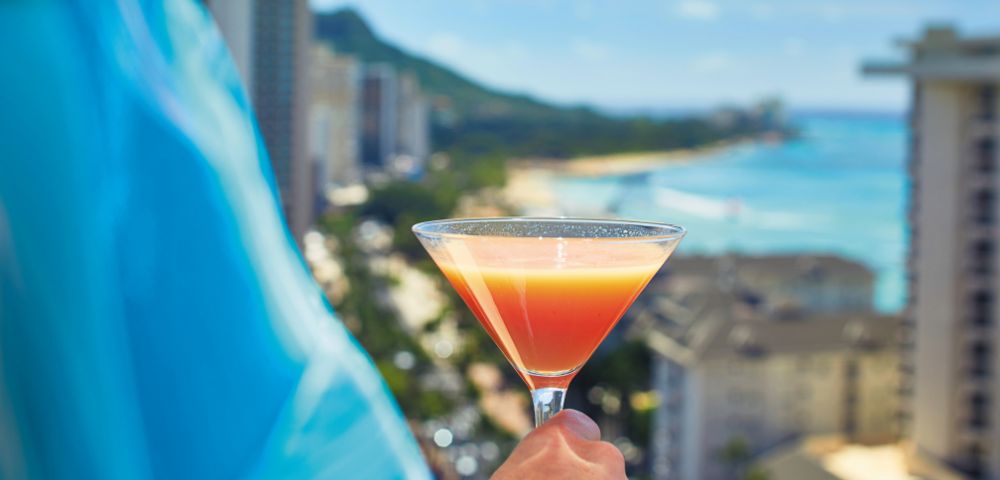A person holds a tropical cocktail in a martini glass, with an ocean view and cityscape in the background. The scene conveys a vacation vibe.
