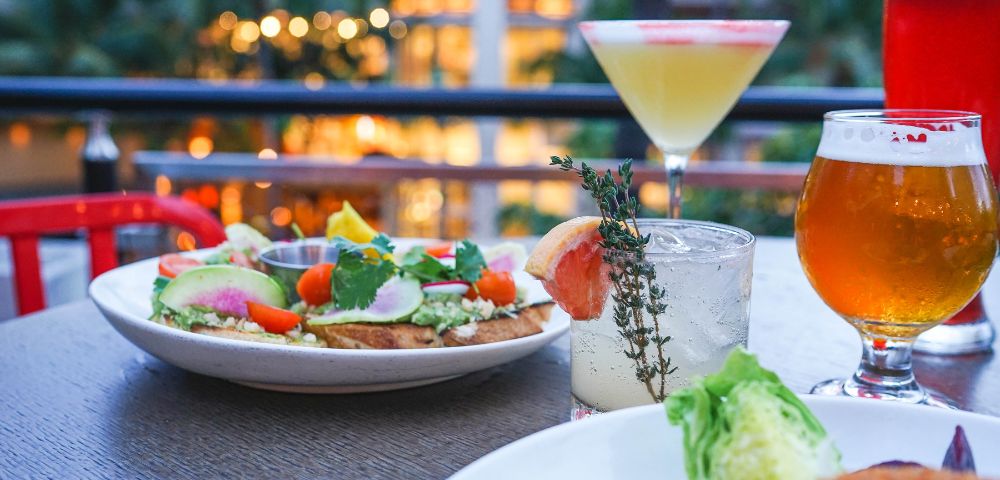 A vibrant outdoor dining scene with a plate of avocado toast and colorful vegetables. Beside it are a cocktail, a craft beer, and a whiskey glass. Warm, inviting ambiance.