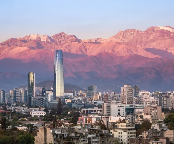 A stunning cityscape set against a dramatic natural backdrop. In the foreground, a modern urban skyline features tall buildings, including a prominent glass skyscraper that stands out as the tallest structure. Surrounding it are other contemporary high-rises and residential buildings. In the background, the majestic Andes Mountains dominate the scene, bathed in warm pink and purple hues from the setting or rising sun, with snow-capped peaks visible at the highest elevations.