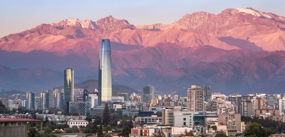A stunning cityscape set against a dramatic natural backdrop. In the foreground, a modern urban skyline features tall buildings, including a prominent glass skyscraper that stands out as the tallest structure. Surrounding it are other contemporary high-rises and residential buildings. In the background, the majestic Andes Mountains dominate the scene, bathed in warm pink and purple hues from the setting or rising sun, with snow-capped peaks visible at the highest elevations.