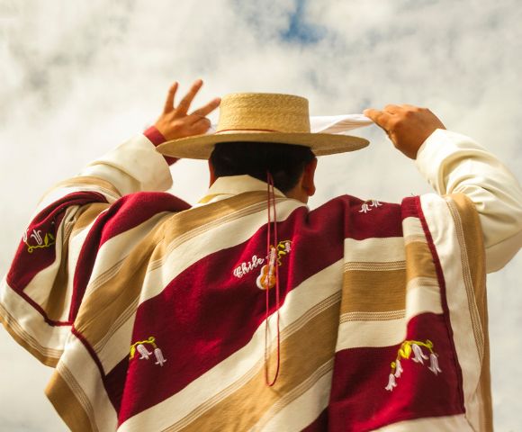 A person wearing a traditional Chilean poncho and straw hat holds a white handkerchief aloft, likely performing a cultural dance under a cloudy sky.