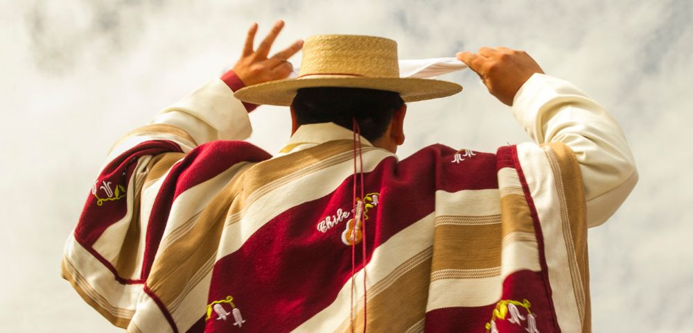 A person wearing a traditional Chilean poncho and straw hat holds a white handkerchief aloft, likely performing a cultural dance under a cloudy sky.