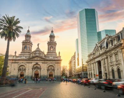 A vibrant cityscape blending historical and modern architecture. In the foreground, there is a grand neoclassical cathedral with ornate details, twin towers, and statues adorning its façade.