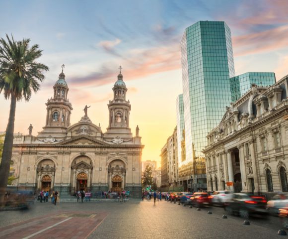 A vibrant cityscape blending historical and modern architecture. In the foreground, there is a grand neoclassical cathedral with ornate details, twin towers, and statues adorning its façade.