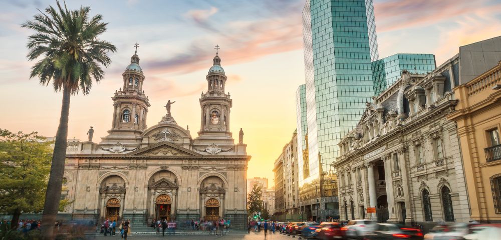 A vibrant cityscape blending historical and modern architecture. In the foreground, there is a grand neoclassical cathedral with ornate details, twin towers, and statues adorning its façade.