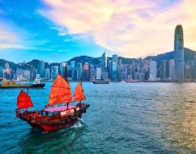 A traditional red-sailed junk boat glides across a vibrant harbor, framed by the modern skyline of skyscrapers under a pastel sunset, evoking a blend of history and modernity.