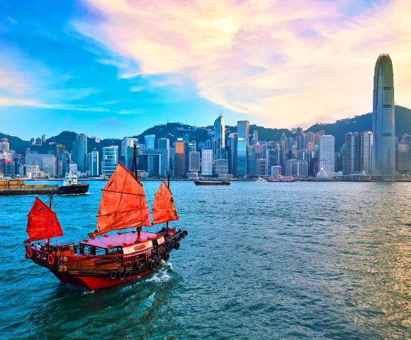 A traditional red-sailed junk boat glides across a vibrant harbor, framed by the modern skyline of skyscrapers under a pastel sunset, evoking a blend of history and modernity.
