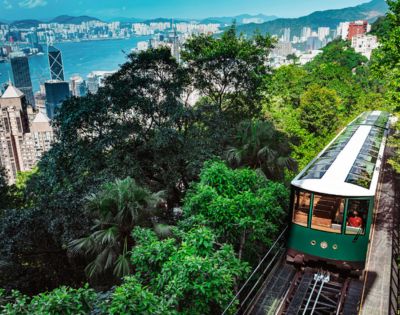 A historic green tram ascends a lush, forested hill overlooking Hong Kong's skyline and harbor, under a bright, clear sky, evoking a sense of adventure.