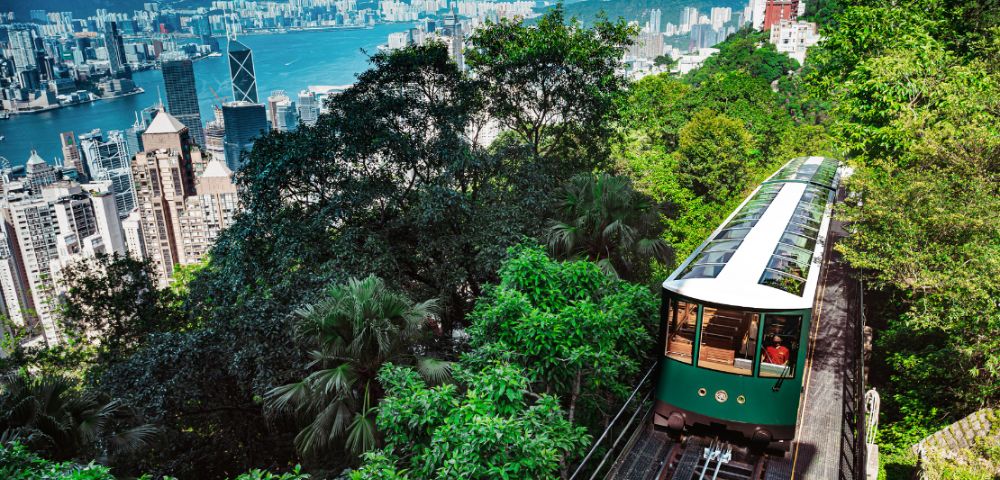 A historic green tram ascends a lush, forested hill overlooking Hong Kong's skyline and harbor, under a bright, clear sky, evoking a sense of adventure.