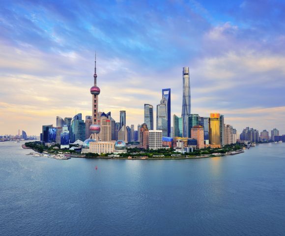 Shanghai skyline at dusk, showcasing iconic skyscrapers like the Oriental Pearl Tower. Calm river in foreground, under a sky painted with soft clouds.