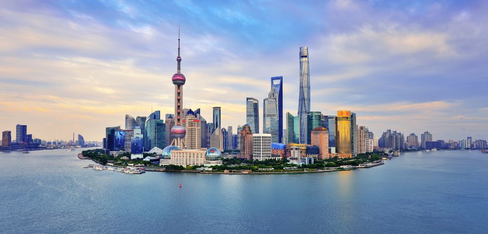 Shanghai skyline at dusk, showcasing iconic skyscrapers like the Oriental Pearl Tower. Calm river in foreground, under a sky painted with soft clouds.