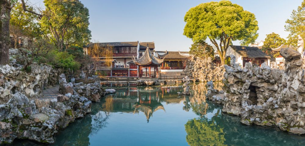Scenic garden with a tranquil pond reflecting a traditional Chinese pavilion and lush trees. Rocky formations and historic buildings surround the water.