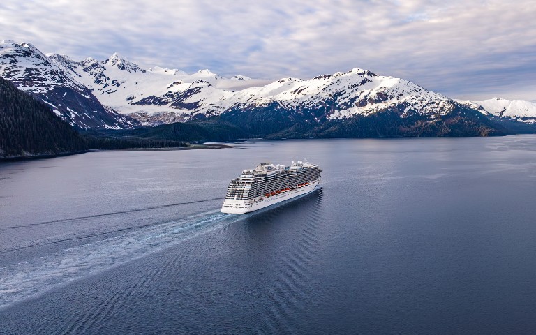 A large cruise ship sails through calm blue waters, leaving a gentle wake. Snow-capped mountains rise under a cloudy sky, creating a serene, majestic scene.