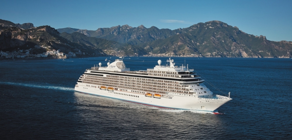 Cruise ship sails on a calm blue sea with a backdrop of rugged mountains under a clear sky, conveying a serene and majestic travel experience.