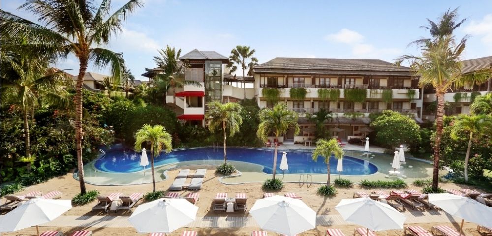Tropical resort scene with a serene pool, surrounded by palm trees and a hotel building in the background. Lounge chairs and umbrellas on a sandy area in the foreground.