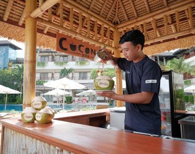 A man pours a drink into a coconut at an open-air tropical bar, with palm trees and umbrellas in the background, conveying a relaxed, vacation vibe.