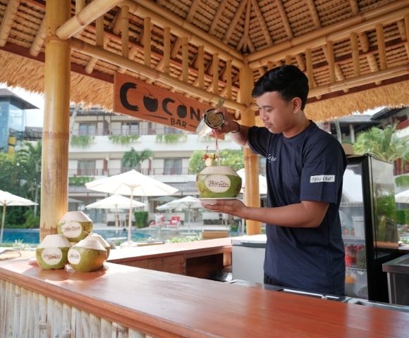 A man pours a drink into a coconut at an open-air tropical bar, with palm trees and umbrellas in the background, conveying a relaxed, vacation vibe.