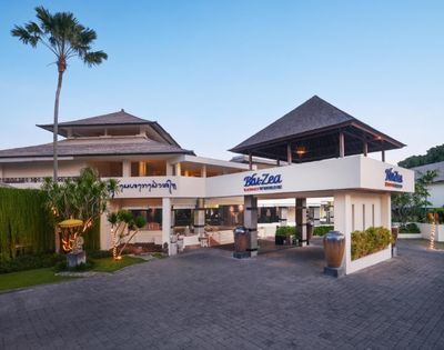 A modern building with Balinese architecture features a peaked roof, tropical plants, and signs reading 