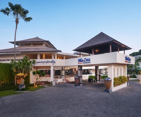 A modern building with Balinese architecture features a peaked roof, tropical plants, and signs reading 