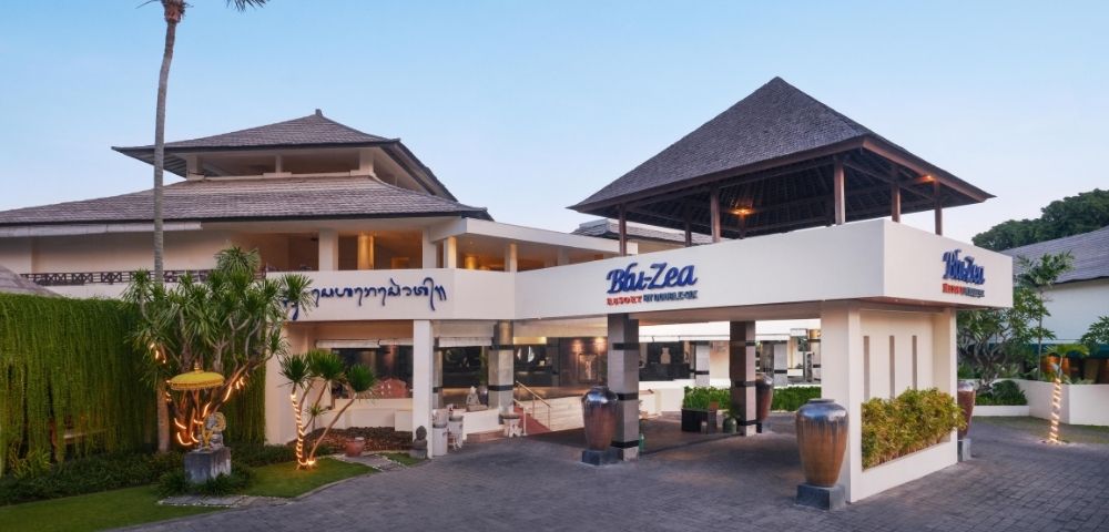 A modern building with Balinese architecture features a peaked roof, tropical plants, and signs reading 