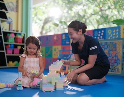 A young girl and a woman sit on a blue mat in a colorful playroom, joyfully building with pastel blocks. Bright light filters through a window.