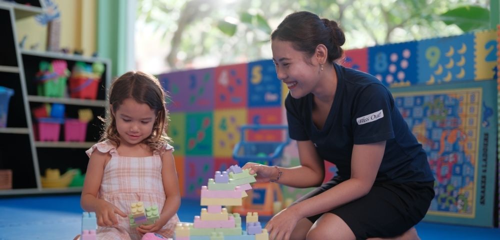 A young girl and a woman sit on a blue mat in a colorful playroom, joyfully building with pastel blocks. Bright light filters through a window.