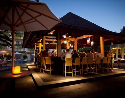 Evening poolside bar scene with people seated on bar stools under a wooden gazebo. Warm lighting and lanterns create a relaxed atmosphere.