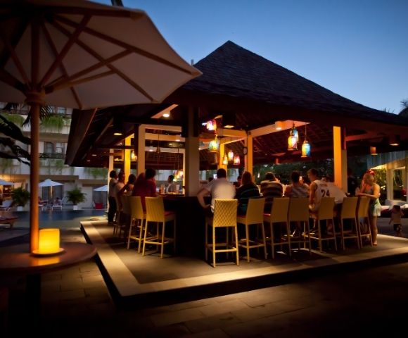 Evening poolside bar scene with people seated on bar stools under a wooden gazebo. Warm lighting and lanterns create a relaxed atmosphere.