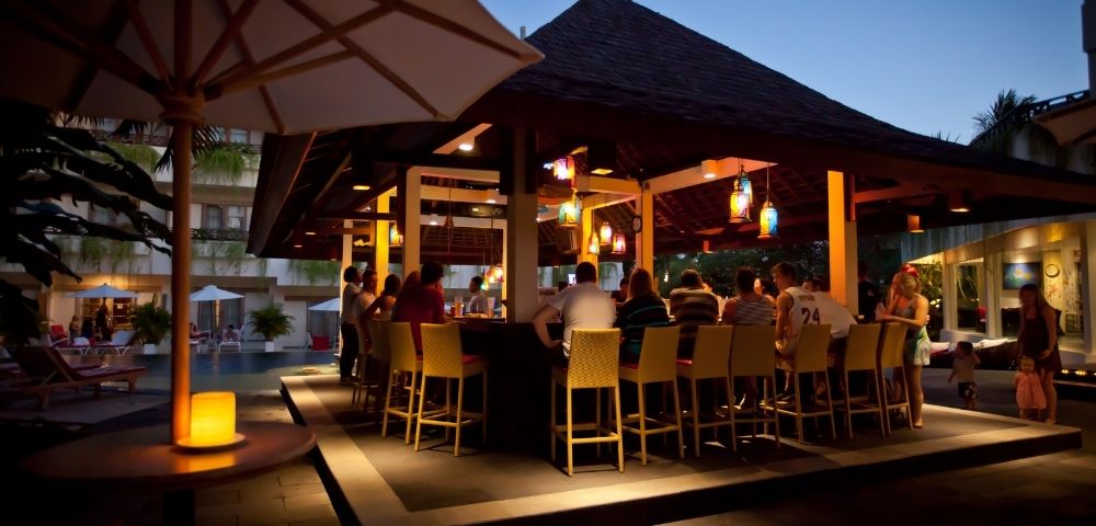 Evening poolside bar scene with people seated on bar stools under a wooden gazebo. Warm lighting and lanterns create a relaxed atmosphere.
