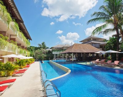 Luxurious resort pool area with bright blue water, surrounded by red lounge chairs, lush greenery, and a tropical vibe under a clear sky with fluffy clouds.