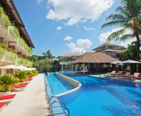 Luxurious resort pool area with bright blue water, surrounded by red lounge chairs, lush greenery, and a tropical vibe under a clear sky with fluffy clouds.