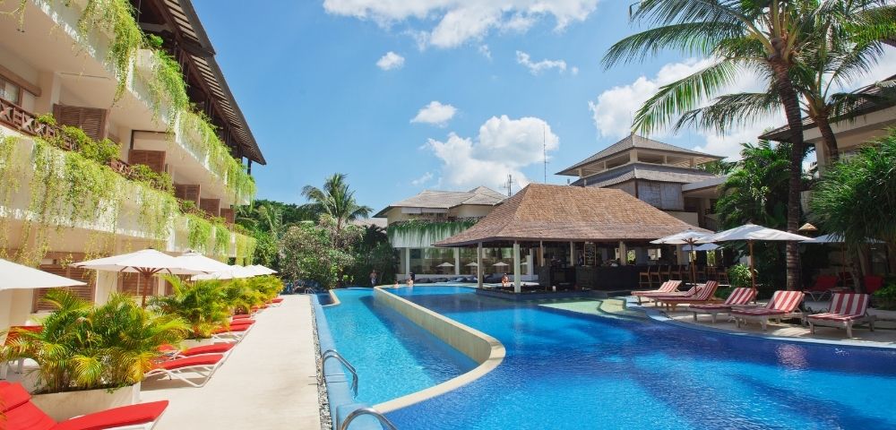 Luxurious resort pool area with bright blue water, surrounded by red lounge chairs, lush greenery, and a tropical vibe under a clear sky with fluffy clouds.