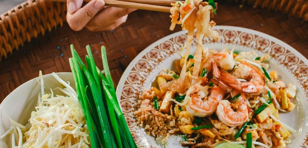 Thai food served with chopsticks, featuring colourful dishes like pad thai and sprouts on a wooden table.