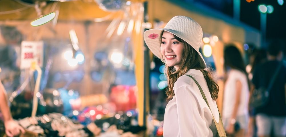 Asian woman wearing a white hat, smiling at a vibrant night market filled with colorful stalls and lights.