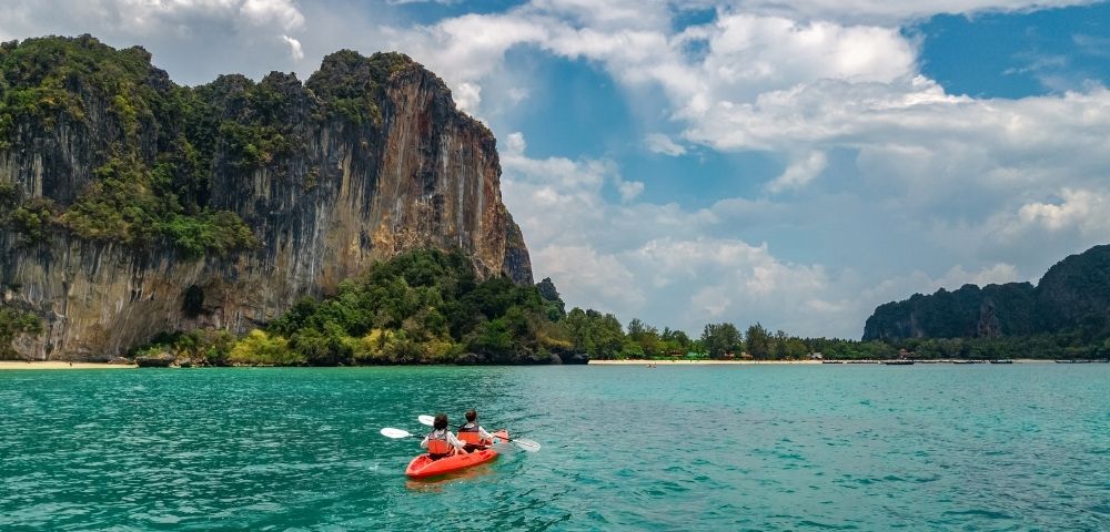 Two people paddle a kayak on water near a rocky island under a clear blue sky.