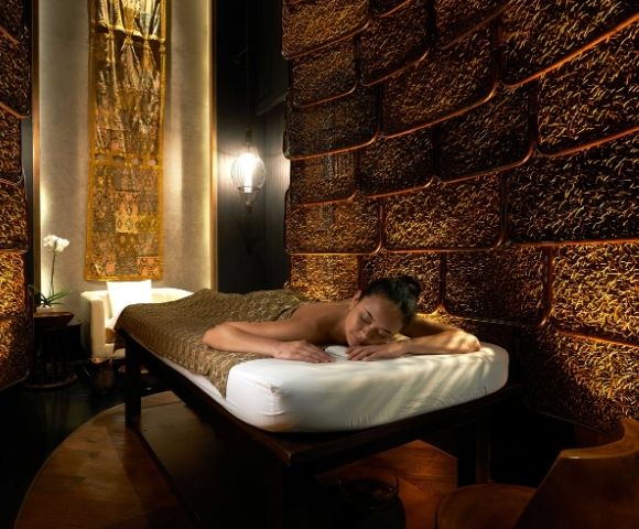 A woman relaxes on a massage table in a dimly lit spa with textured brown walls and warm lighting. The atmosphere is calm and luxurious.