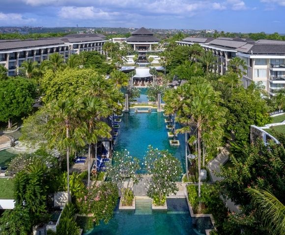 Aerial view of a luxurious resort with a long, central pool lined by lush palm trees and surrounded by elegant buildings, conveying a serene, tropical ambiance.