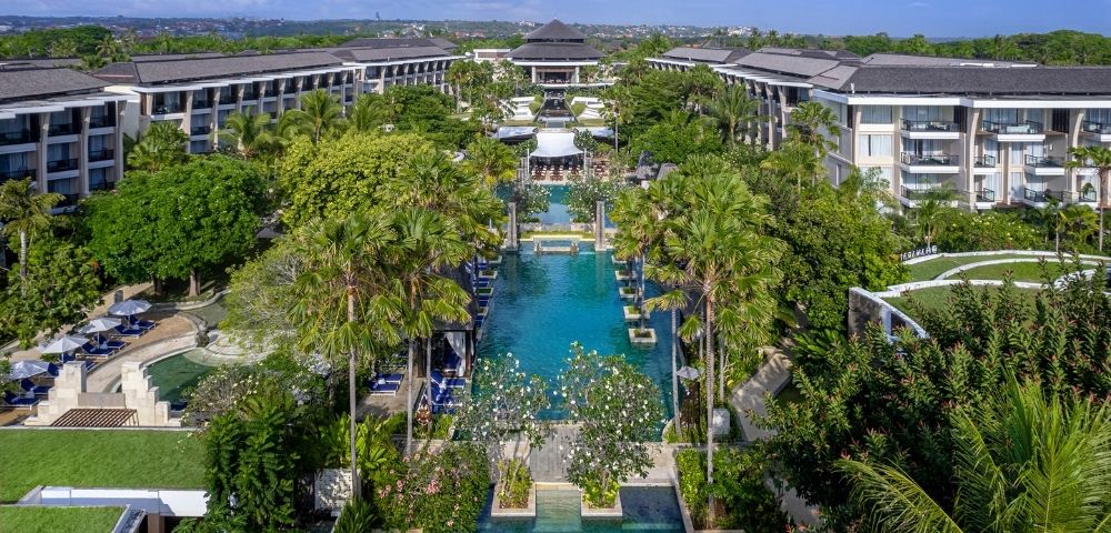 Aerial view of a luxurious resort with a long, central pool lined by lush palm trees and surrounded by elegant buildings, conveying a serene, tropical ambiance.