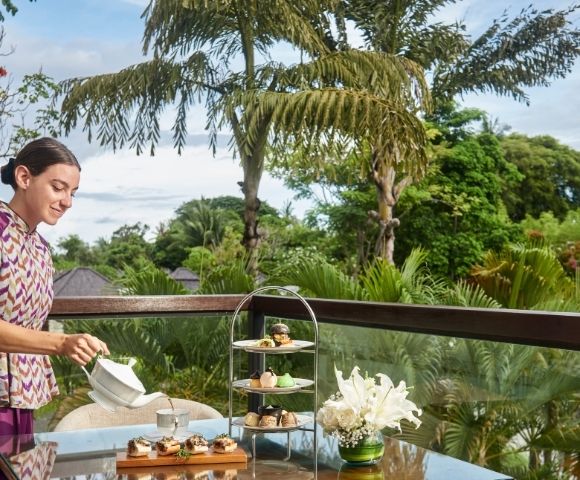A woman in a patterned dress pours tea at an outdoor table. The table features a tiered tray of treats and flowers. The background shows lush greenery and a blue sky.