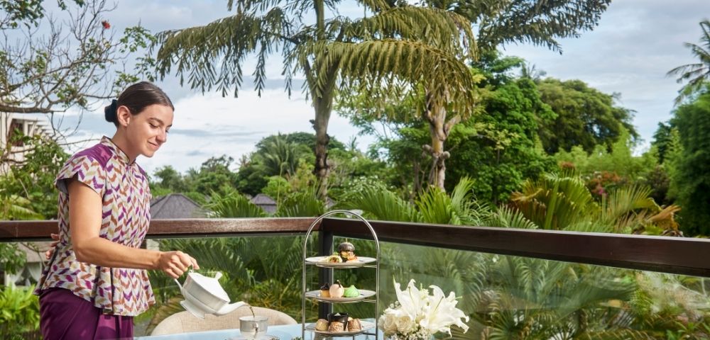 A woman in a patterned dress pours tea at an outdoor table. The table features a tiered tray of treats and flowers. The background shows lush greenery and a blue sky.