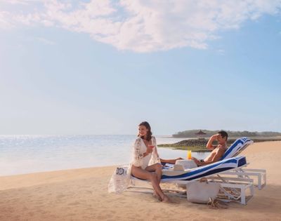 A couple relaxes on beach loungers under a clear sky. The woman sits up smiling, while the man reclines beside her. A calm ocean and sandy beach frame the scene.