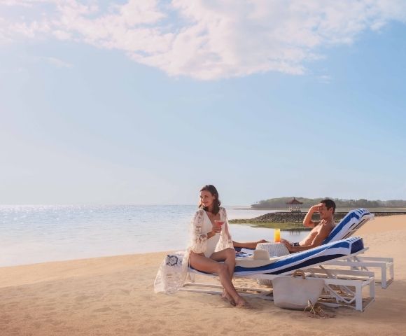 A couple relaxes on beach loungers under a clear sky. The woman sits up smiling, while the man reclines beside her. A calm ocean and sandy beach frame the scene.