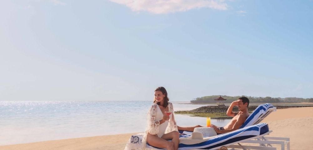 A couple relaxes on beach loungers under a clear sky. The woman sits up smiling, while the man reclines beside her. A calm ocean and sandy beach frame the scene.