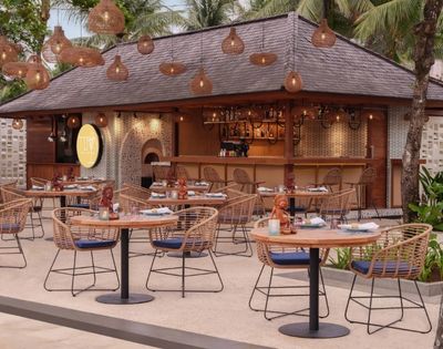 Outdoor tropical restaurant with wicker chairs and round tables set for dining. Warm lighting from hanging lamps and lush greenery create a cozy atmosphere.
