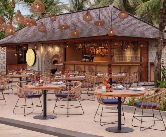Outdoor tropical restaurant with wicker chairs and round tables set for dining. Warm lighting from hanging lamps and lush greenery create a cozy atmosphere.