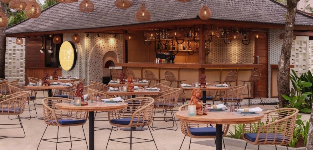 Outdoor tropical restaurant with wicker chairs and round tables set for dining. Warm lighting from hanging lamps and lush greenery create a cozy atmosphere.